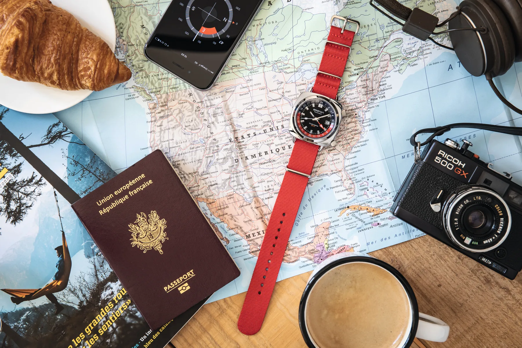 Briston GMT watch on a red NATO strap laid beside a passport, map and espresso cup