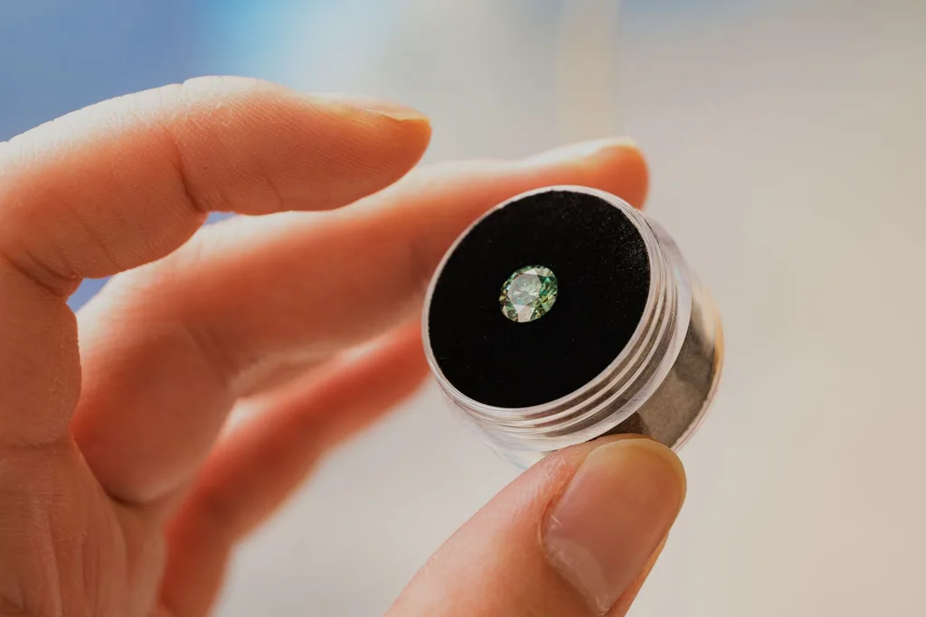 A loose green gemstone in a display jar held between 2 fingers