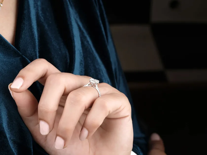 An elongated diamond engagement ring on a hand against teal velvet