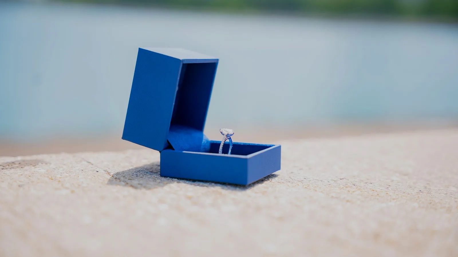 A diamond engagement ring in an open blue box on a sunlit stone ledge