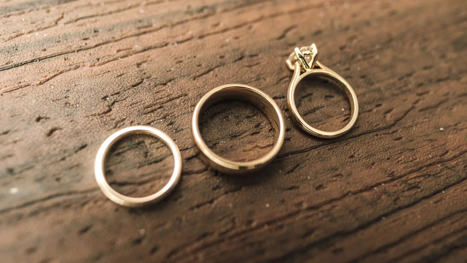 Three gold rings on dark wood, a thin band, a wide band and a diamond solitaire