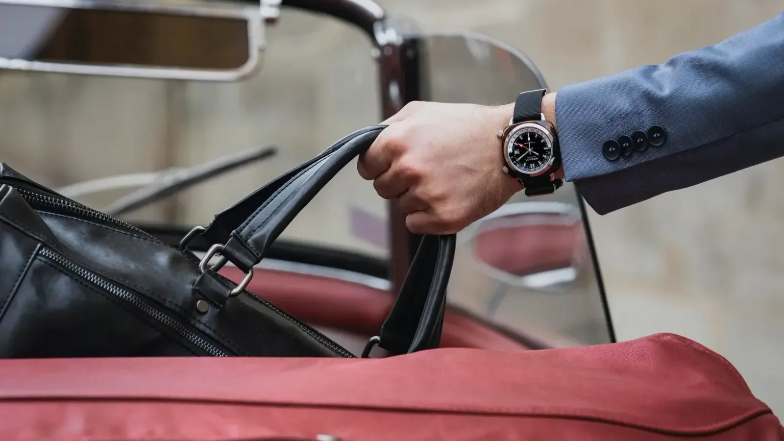 A wristwatch worn at the cuff as a hand lifts a leather bag into a car