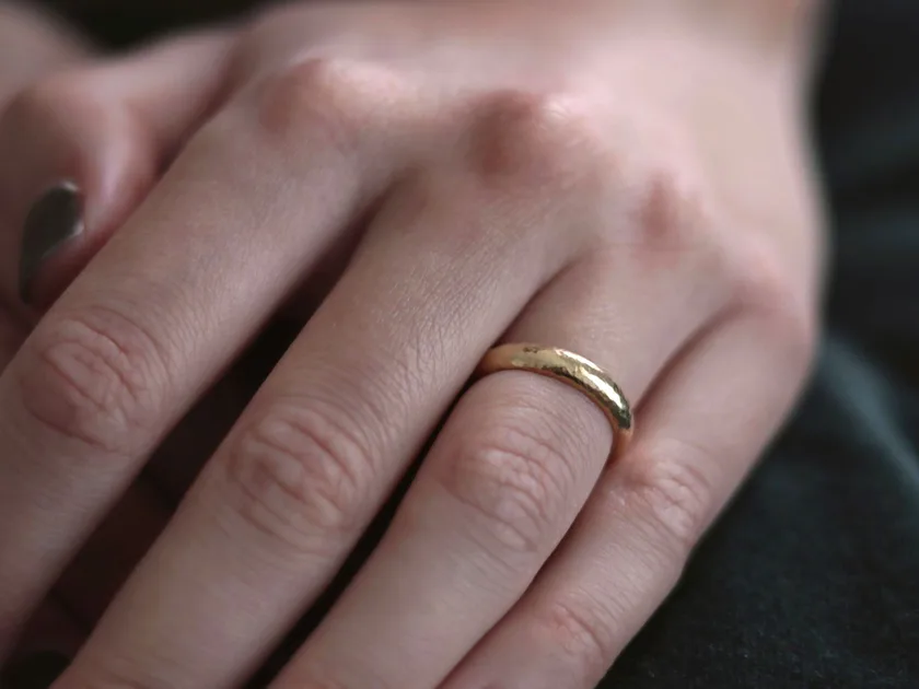 A plain hammered gold band on a hand against a dark background