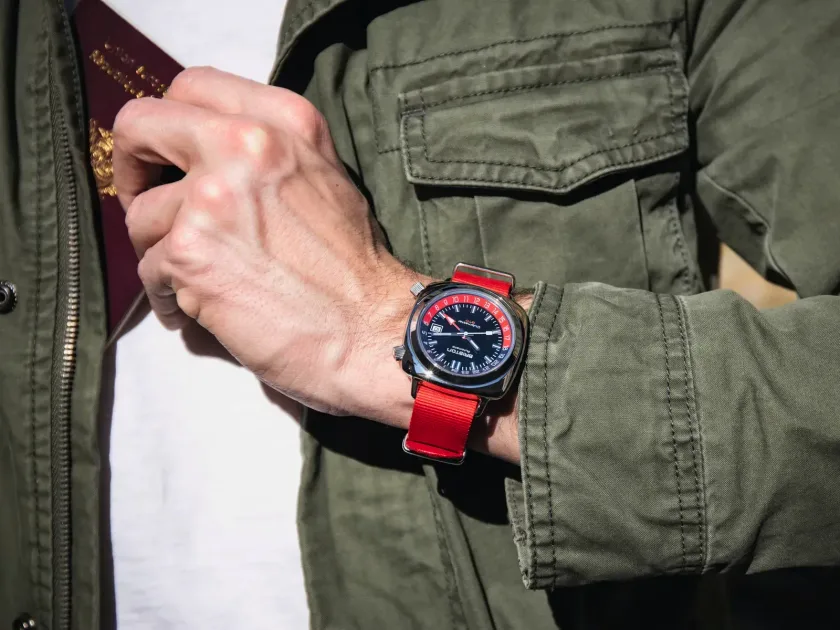 A Briston watch on a red NATO strap worn with an olive field jacket