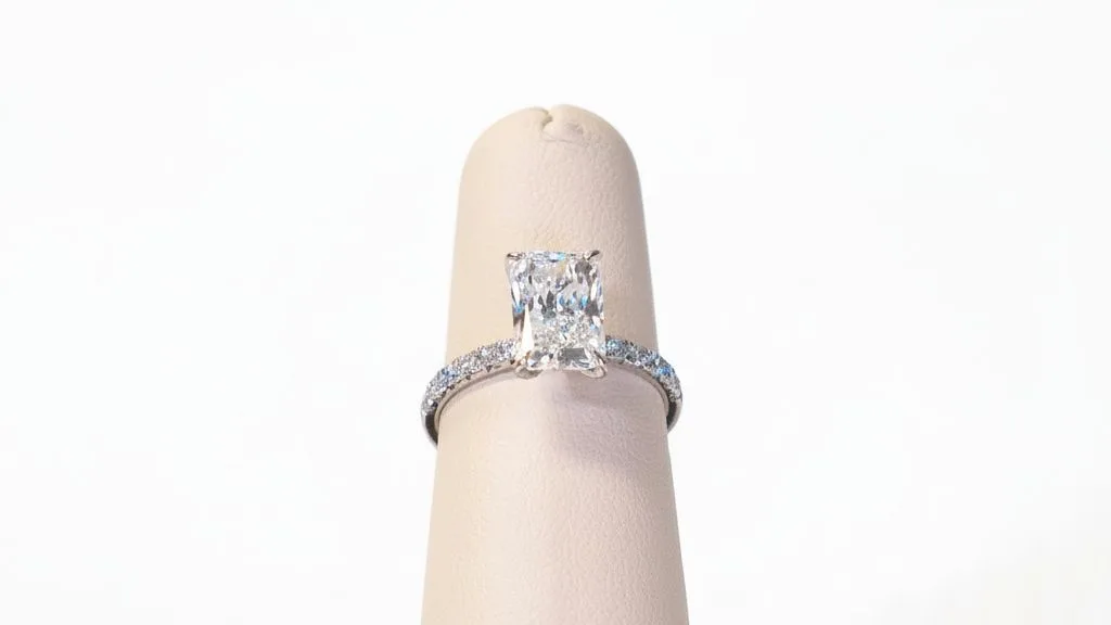 A cushion cut diamond solitaire held by prongs above a pave set band